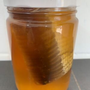 Frasco de mel transparente contendo um favo de abelha dentro, colocado sobre uma superfície escura.